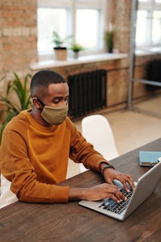 Adult in face mask typing on laptop indoors near large windows. Bright natural light setting.
