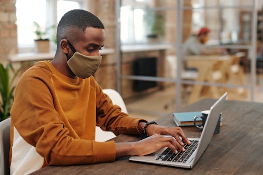 Man wearing a facemask typing on a laptop indoors with blurred background.