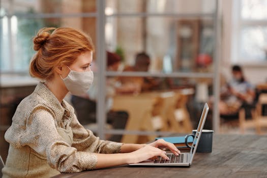 Red-haired woman wearing mask, working on laptop in trendy indoor office space.