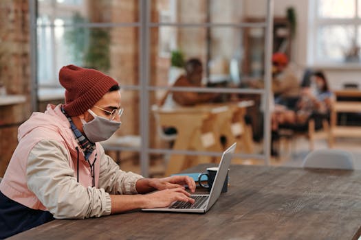 A young man in a beanie and mask typing on a laptop at a wooden table indoors.