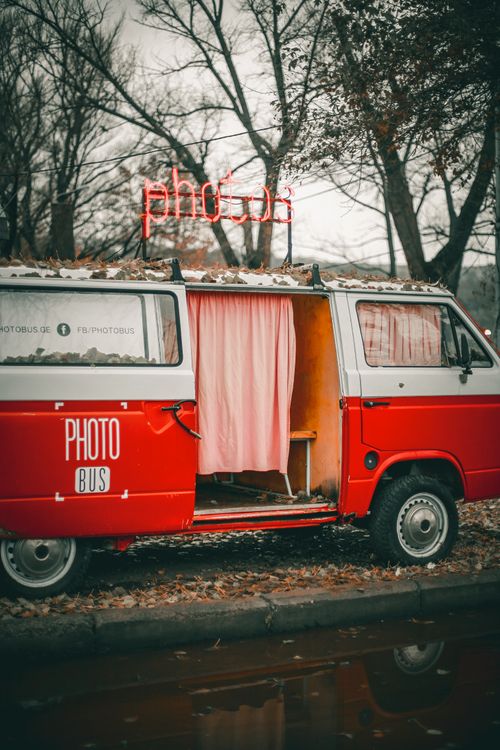 Free Red retro photo bus van with opened door with curtains parked on ground with fallen leaves near asphalt road with puddle and trees in daytime under gray sky in countryside Stock Photo