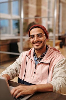 A young man wearing a beanie and glasses, smiling while using a laptop in a stylish open-plan office.