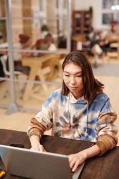 Focused young woman in hoodie using laptop at modern coworking space, engaged in work.