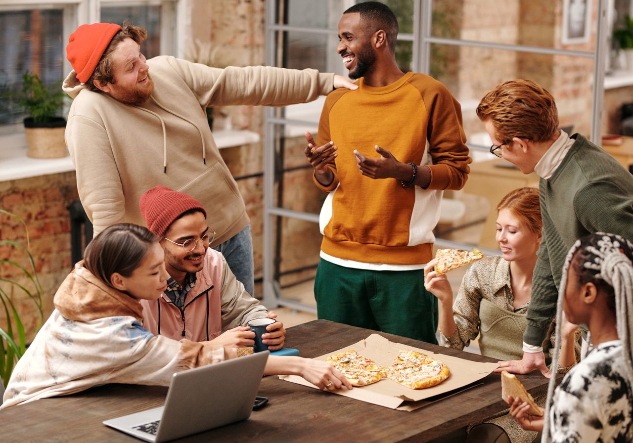 A diverse group of adult friends laughing and sharing pizza around a table indoors