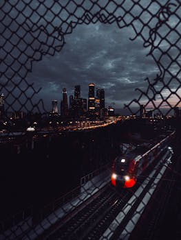 A dynamic night cityscape featuring a moving train and illuminated skyscrapers through a chain-link fence.
