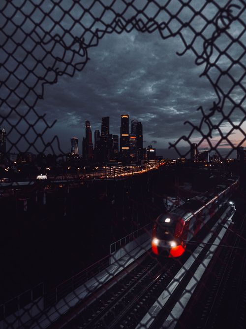 Free A dynamic night cityscape featuring a moving train and illuminated skyscrapers through a chain-link fence. Stock Photo