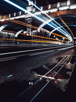 Stunning long exposure at night capturing vehicle light trails reflecting in puddles under an urban bridge.