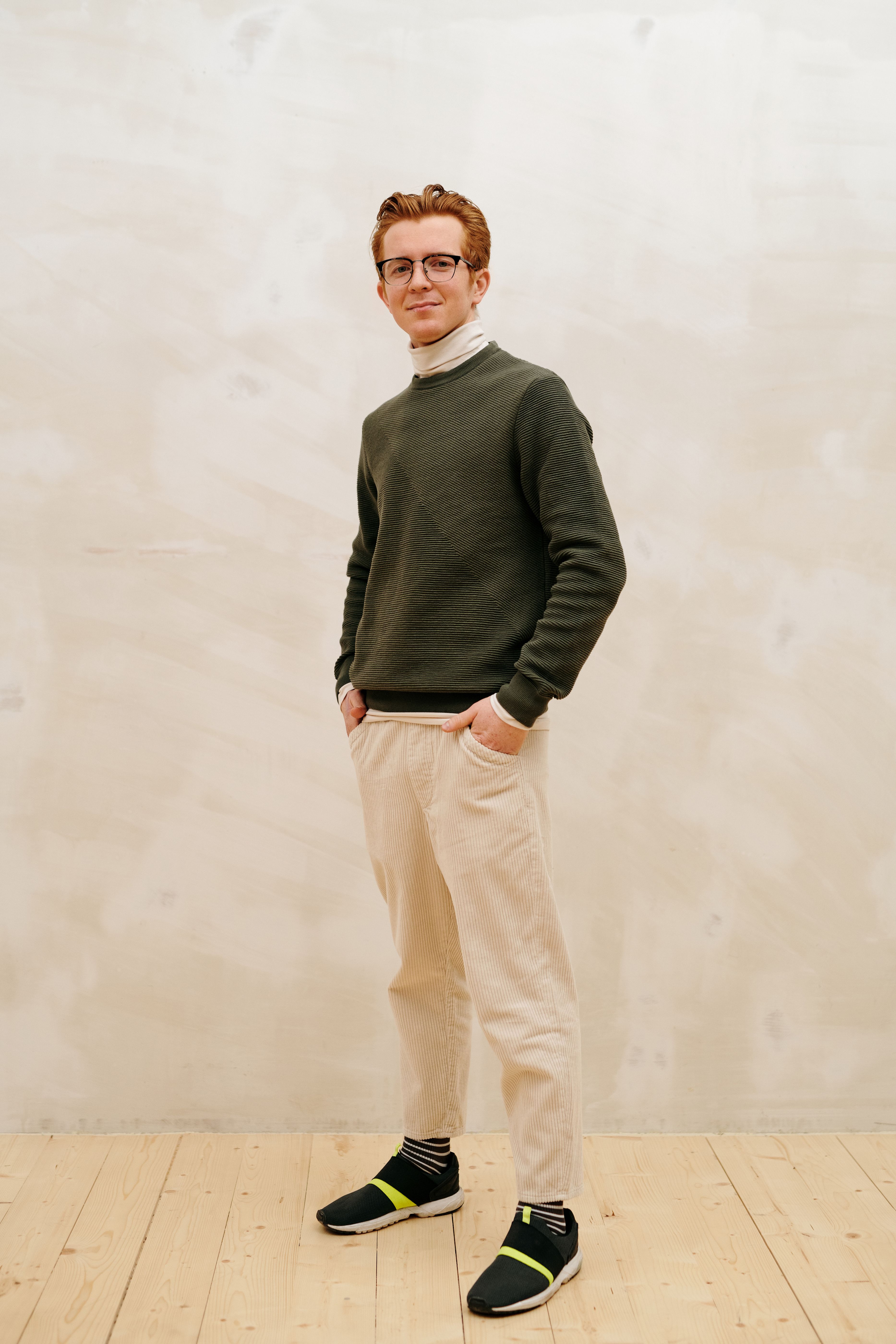 Smiling man in green sweater and beige pants, hands in pockets, indoors.