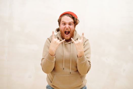 A bearded man in a beige hoodie enthusiastically shows a rock hand sign indoors.