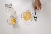 Top view of a bowl of cereal with milk, a spoon, and a milk bottle on a white surface.