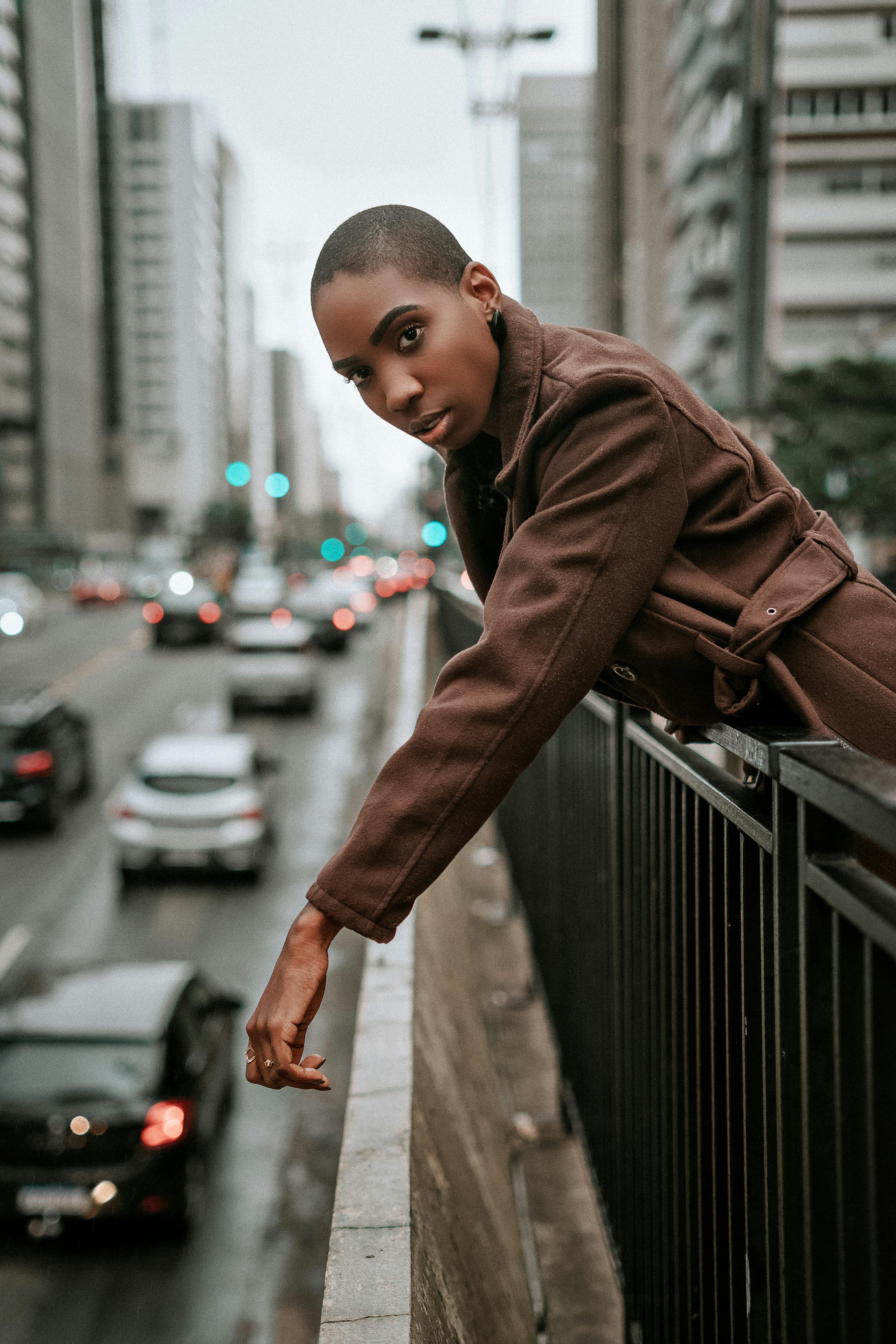 Serious young African American female in brown coat with short hair standing on footbridge over road near metal fence and looking at camera