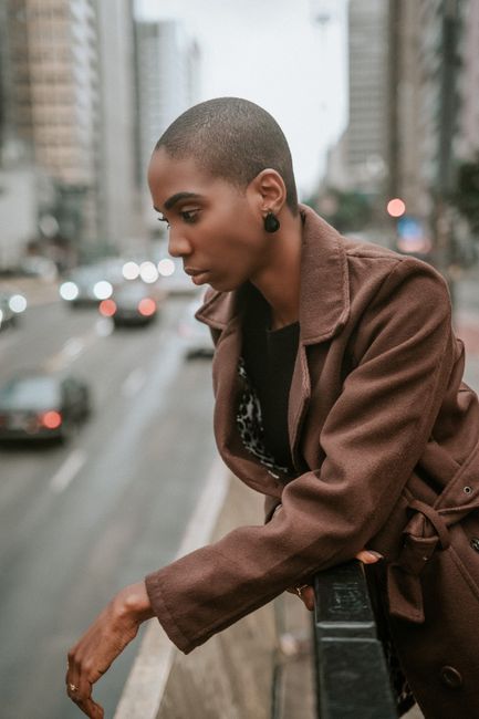 Pensive young African American female with shaved haircut in brown coat standing on footbridge near railing and looking down