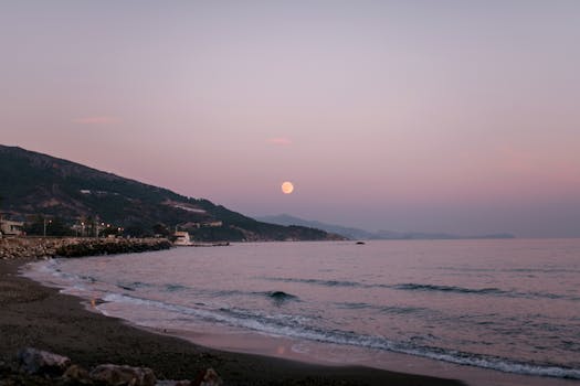 Stunning beach view with a full moon rising over mountains, captured at twilight.