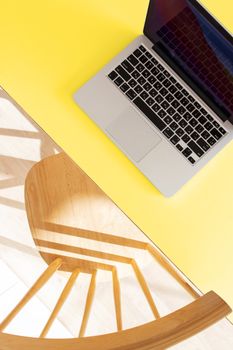 Aerial view of a laptop on a vibrant yellow desk with a curved wooden chair casting shadows.