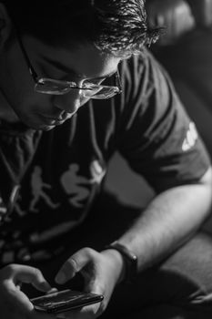 Young man wearing glasses intensely focused on his smartphone in a grayscale setting.