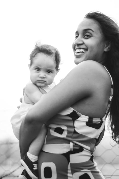Black and white of happy ethnic mother in sundress carrying funny adorable infant and looking away