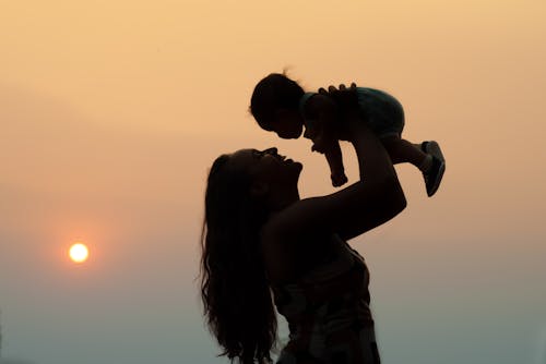 Side view silhouette of loving mother carrying little kid above head standing against sundown sky