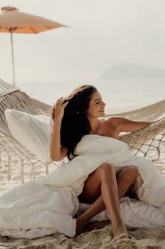 Caucasian woman enjoying a sunny day on a beach hammock in Bali, wrapped in a white blanket.