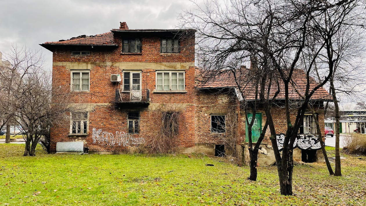 Free A dilapidated brick house with graffiti, surrounded by barren trees on a cloudy day. Stock Photo