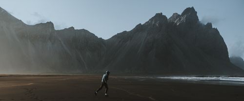 Free Distant unrecognizable tourist in casual clothes walking on empty sandy beach towards mountain range Stock Photo