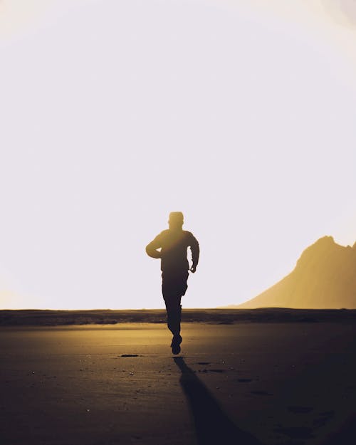 Back view of silhouette of anonymous athletic male running on dark sandy beach at sunset