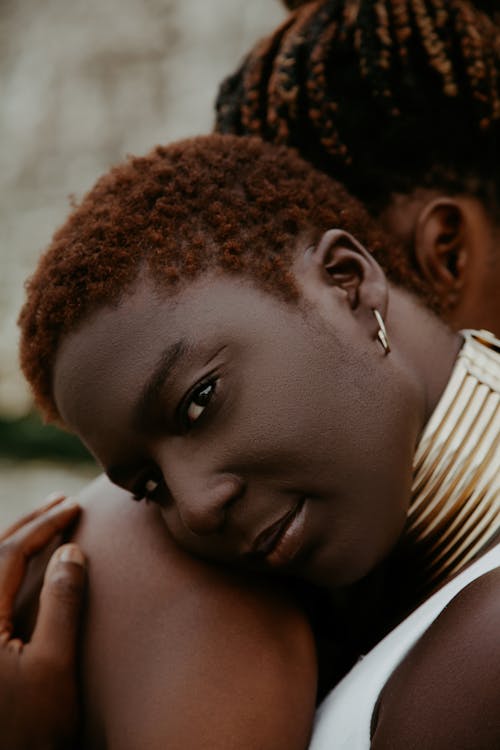 Content young African American female wearing bold golden necklace gently embracing person with bare shoulders and looking at camera