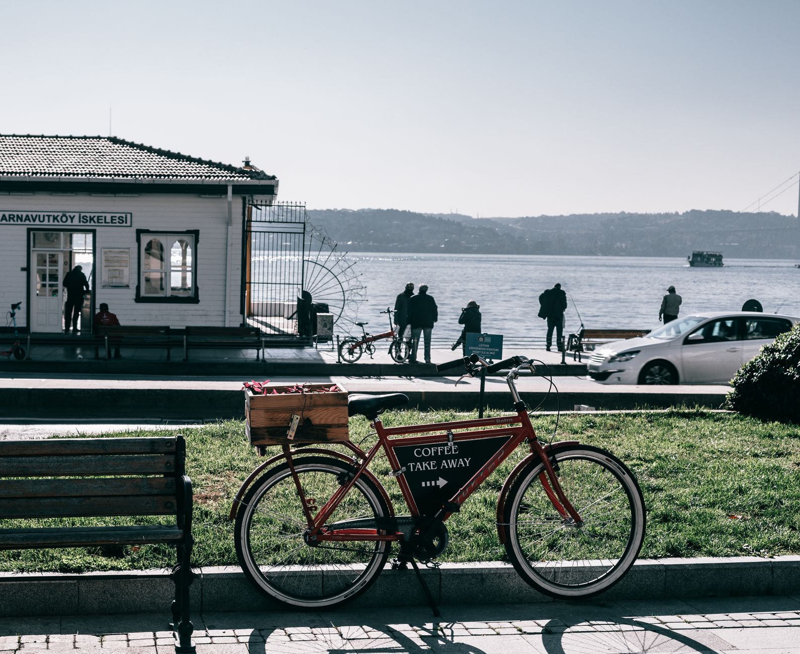 Bicycle with pointer on black board parked on crowded paved embankment with car and building against calm sea and mountain on background