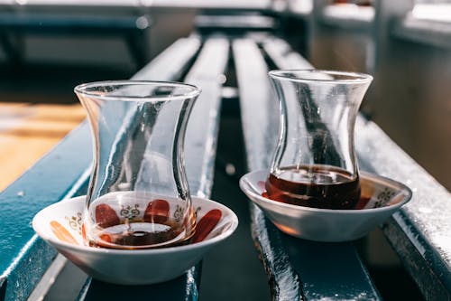 Perspective view of transparent glasses with hot drink served on plates placed on wooden blue bench in room against blurred background