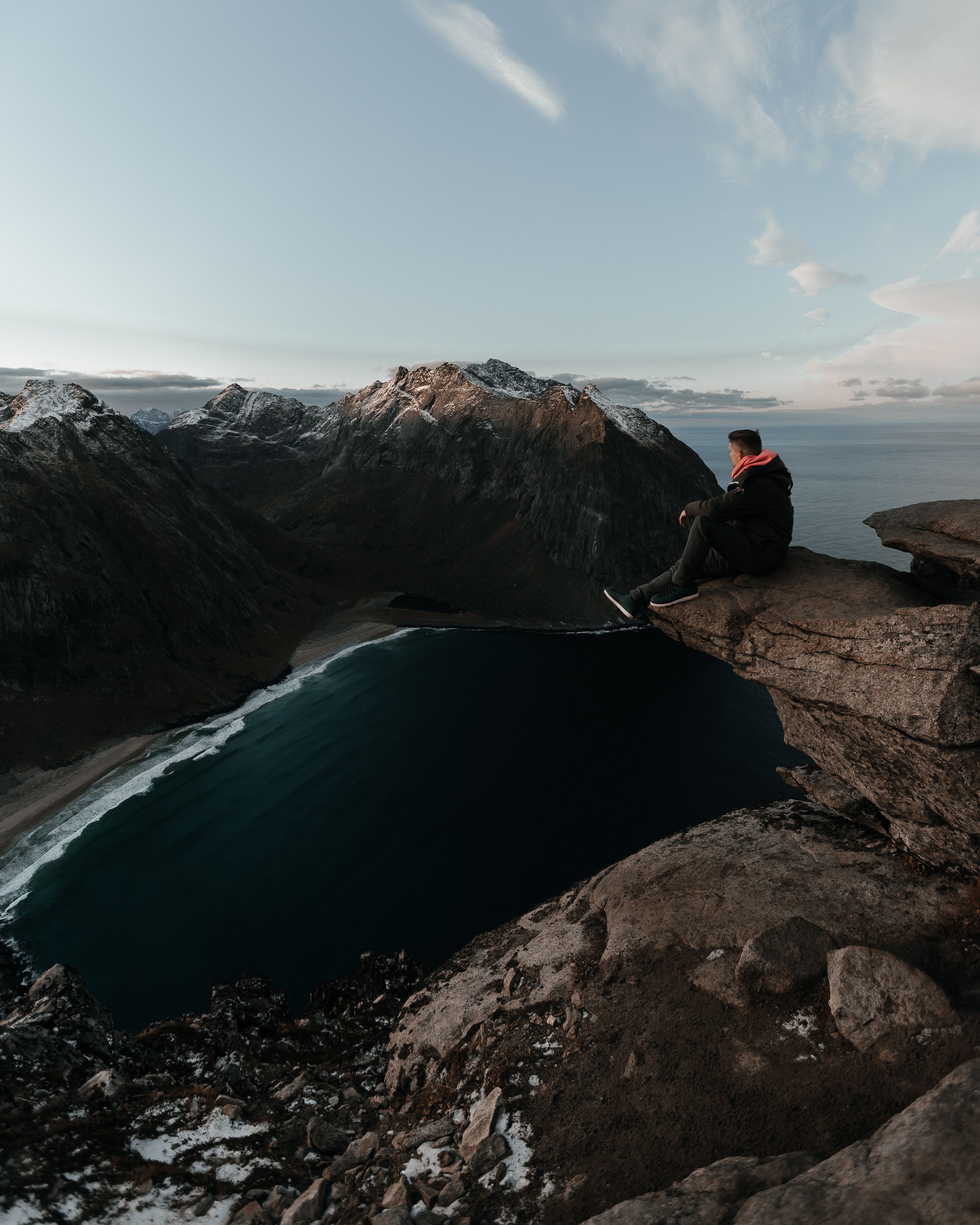 Back view of anonymous traveler in warm clothes sitting on rocky cliff and admiring picturesque scenery of rocky mountains and ocean