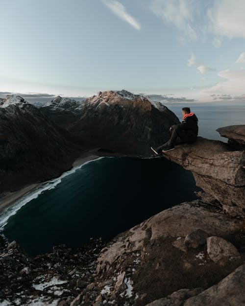Человек в черной куртке сидит на скале возле водоема