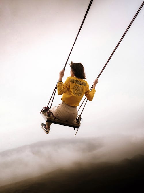 Back view of anonymous female in casual outfit swinging on swing while enjoying view of sky