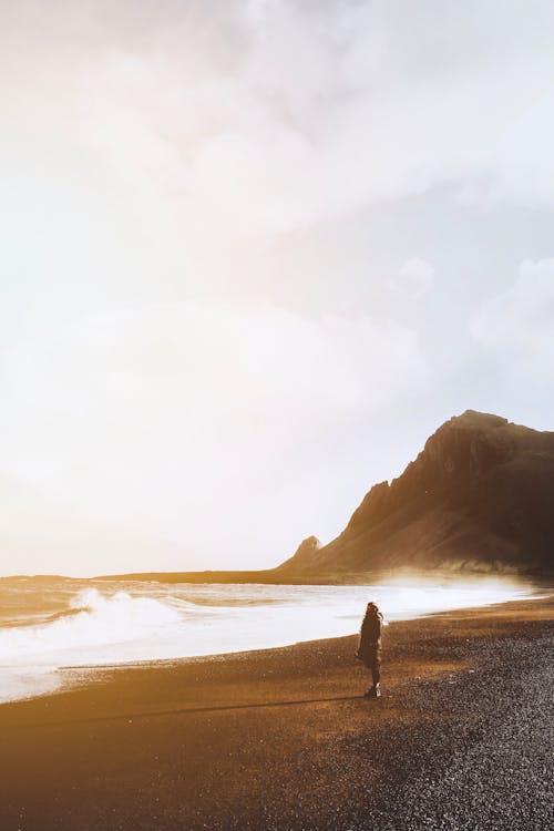 Anonymous distant traveler standing on seashore with rocky cliff while admiring view of stormy sea against cloudy sky in nature