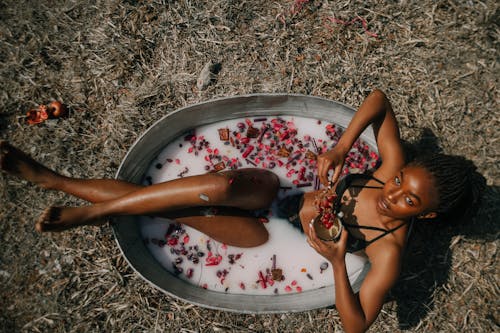 Top view full body of African American female in underwear bathing with flowers and milk in nature