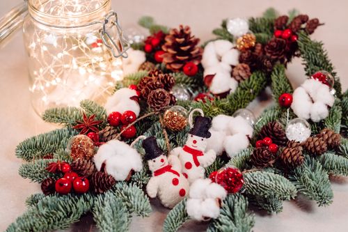 Free Cozy Christmas still life featuring snowman ornaments, pine cones, and twinkling lights on a evergreen wreath. Stock Photo