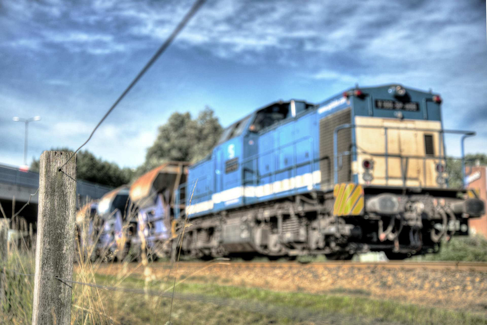 Blurred image of a blue freight train on a railroad track with a wooden fence post in the foreground.