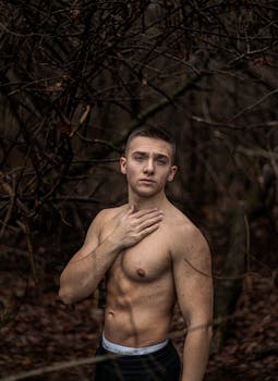 Fit young man with muscular build posing shirtless in a dark, moody forest scene.