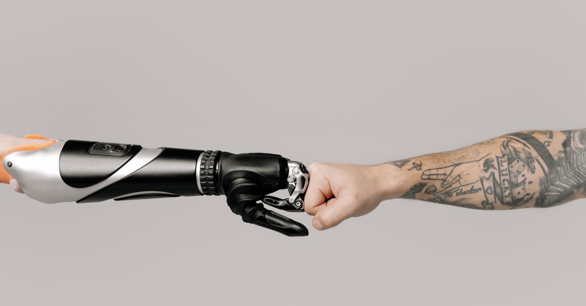 Close-up of a tattooed arm fist bumping with a robotic prosthetic arm in studio setting.