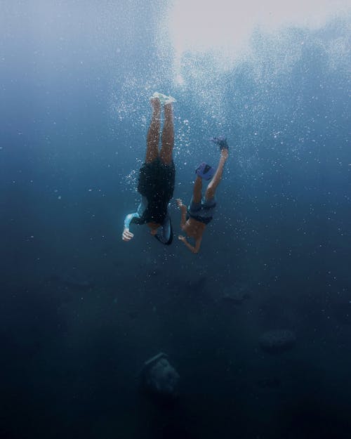 Full body unrecognizable kid and adult person in flippers swimming underwater near bottom with stones