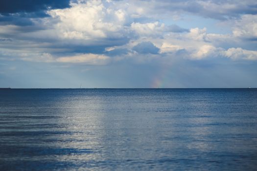 多云天空下，平静的海平线上有一道淡淡的彩虹，营造出宁静的海景