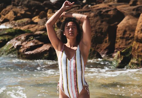 Free Asian woman in striped swimsuit enjoying the beach, arms raised, eyes closed, soaking up the summer sun. Stock Photo