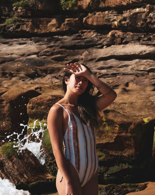 Free A woman in a striped swimsuit stands by a rocky beach, shielding her eyes from the sun. Stock Photo