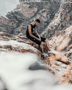 Young man sitting on rocky cliff in a rugged mountain landscape during summer.
