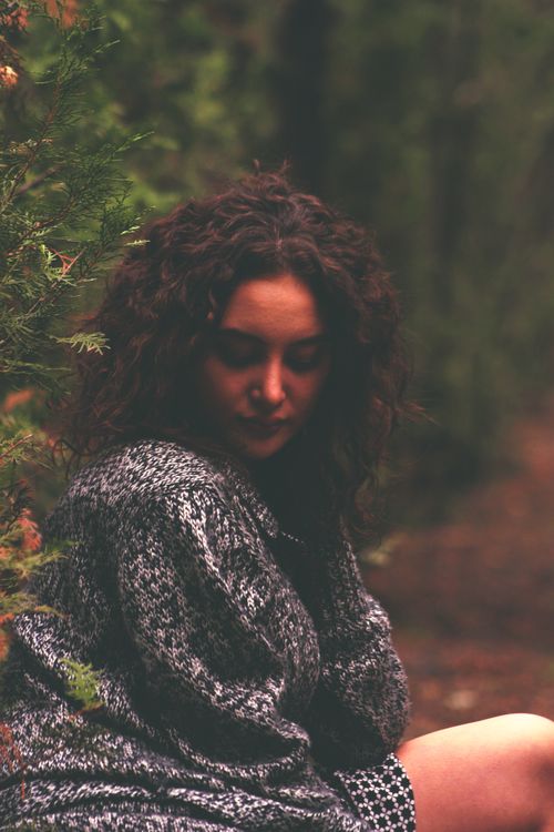 Free A woman with curly hair wearing a knitted sweater sits thoughtfully in a wooded area. Stock Photo