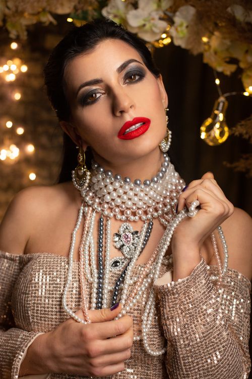 Free Charming lady with bright makeup in expensive pearl necklace and stylish festive outfit standing in room with garlands and decorations and looking at camera Stock Photo