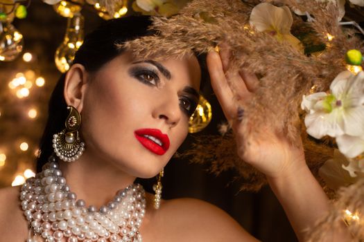 Stylish lady with bright makeup in expensive pearl necklace and earrings standing in room with festive decorations and golden garlands and looking away