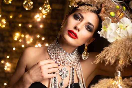 Rich female with dark hair and bright makeup wearing festive pearl necklace and earrings and looking at camera against shiny golden garlands in room with decorations