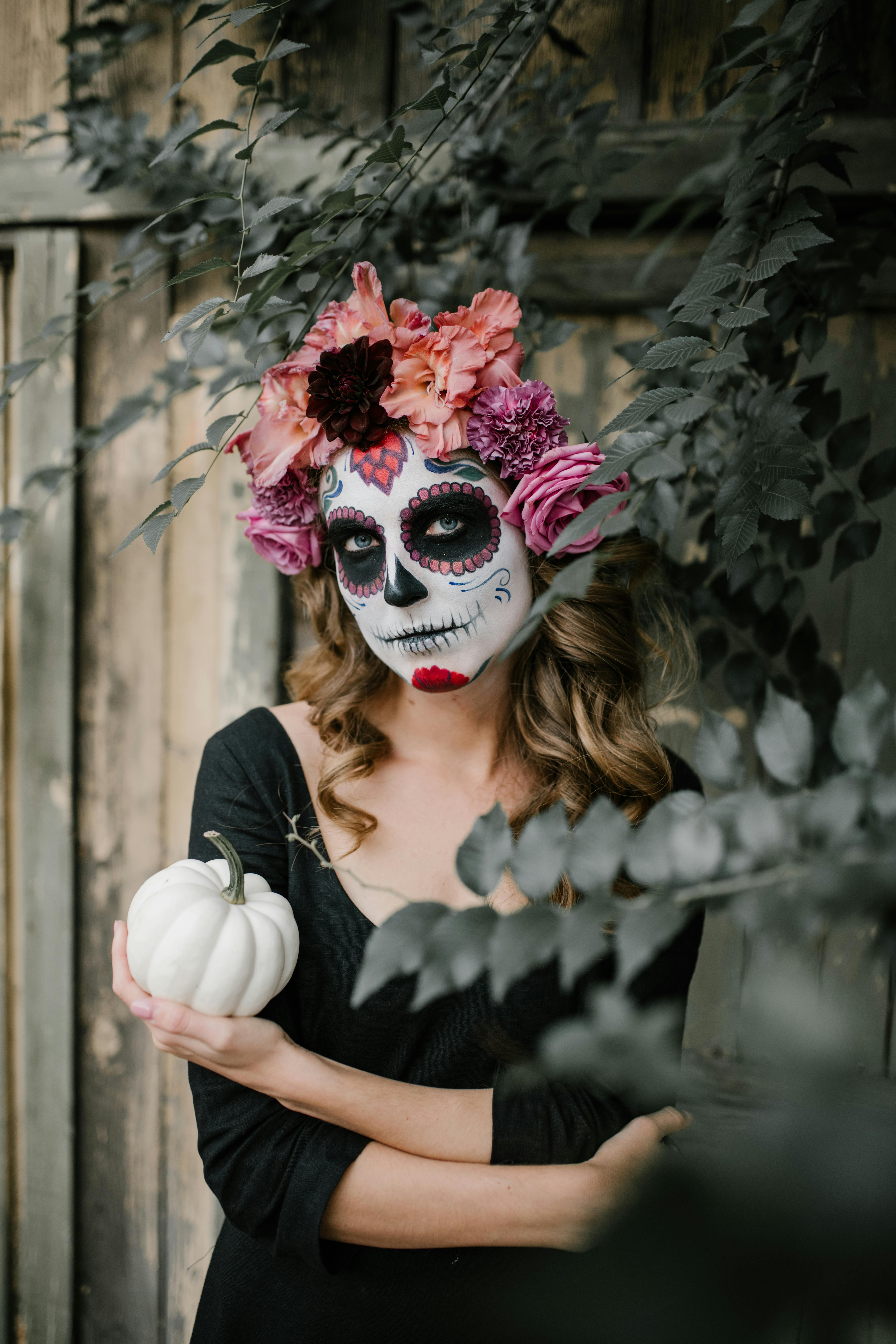 Atmospheric portrait of woman with Dia de Muertos makeup and flower crown holding a small white pumpkin outdoors.