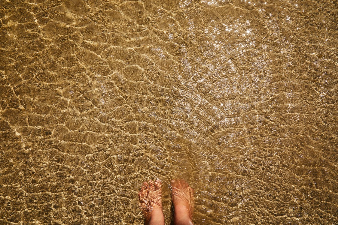 Top view of crop anonymous barefoot female standing in pure transparent rippling water of sea with sandy bottom
