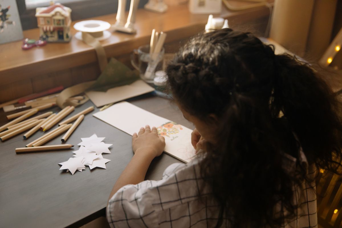 Une enfant de dos, assise à un bureau, décore une carte de vœux entourée de crayons de couleur et d'étoiles en papier découpées