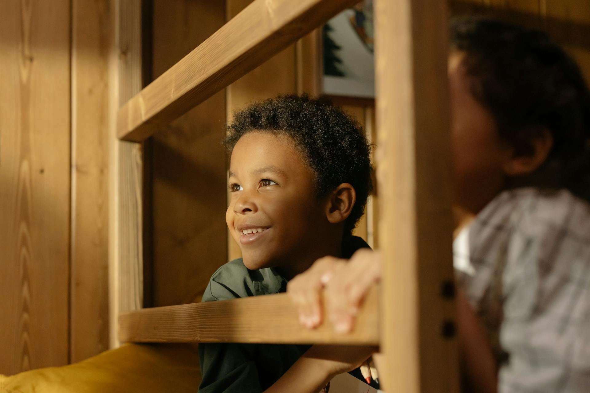 Two cheerful children enjoying playtime on a wooden bunk bed indoors, basking in warm sunlight.